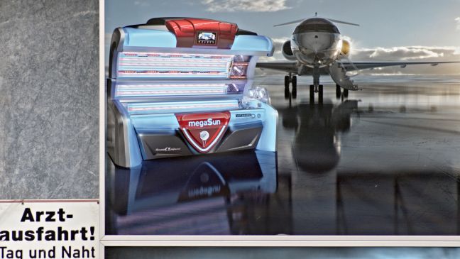 Auf einem Rollfeld steht ein Privatflugzeug, davor eine Sonnenbank, links neben dem Bild ein Schild worauf Arztausfahrt! steht