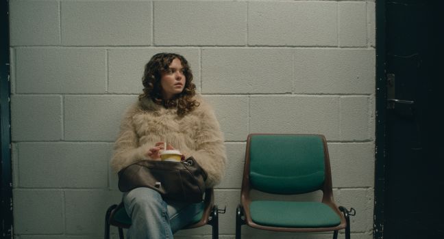 Nora wartend mit Softdrink auf einem Stuhl im Flur