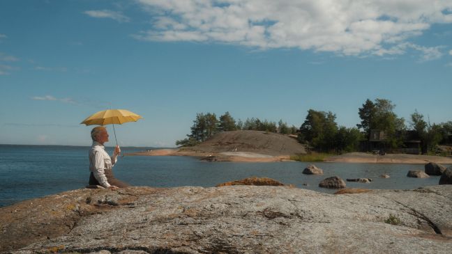 Großmutter sitzt am Strand (Steinfelsen) mit gelbem Schirm