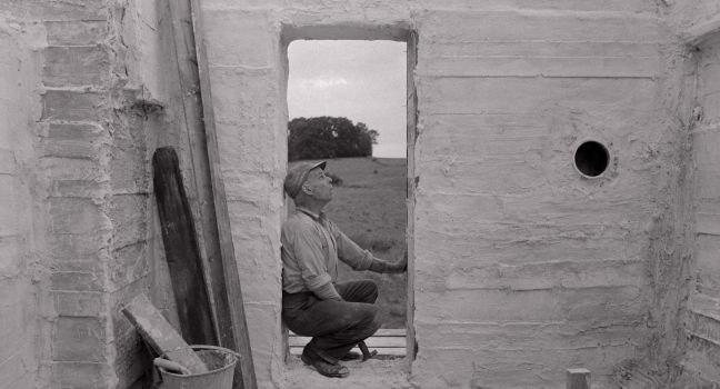 Karl-Göran im Rohbau des Gebäudes mit Hammer in der Hand hockt im Türspalt