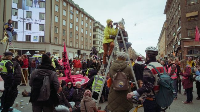 Demonstranten auf der Kreuzung, Dialogpolizisten versuchen ein magentafarbiges Boot zu entfernen, davor zwei Frauen auf einer Leiter mit Megafon