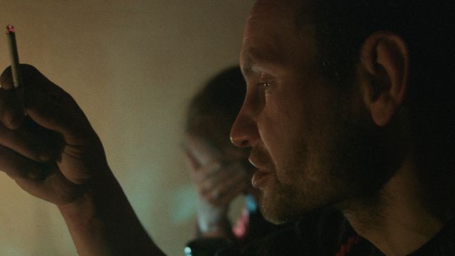 Nahaufnahme eines Mannes mit Zigarette in der Hand, im Hintergrund vergräbt eine Frau ihr Gesicht in den Händen