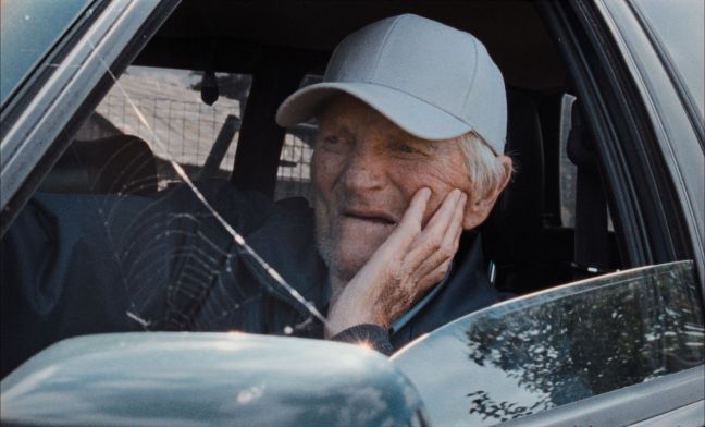 Ein älterer Mann schaut aus dem offenen Fenster seines Autos, eine Spinnwebe zwischen Außenspiegel und A-Säule