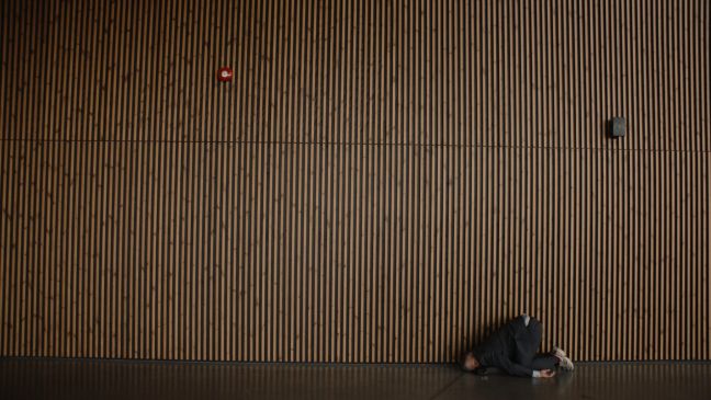 Vor einer großen Wand mit Holzpaneelen, liegt der Mann mit Kopf voran und angezogenen Beinen