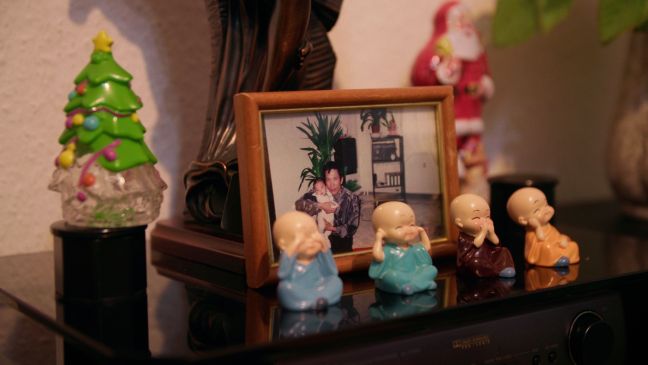 Hung mit Anna als Baby auf dem Arm auf einem Foto, davor vier kleine Buddhas, Weihnachtsdekoration im Hintergrund