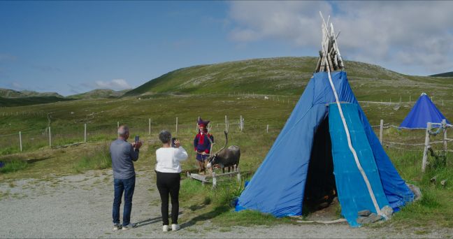 Ein Sámi posiert in Tracht mit einem Rentier neben einem blauen Zelt für die Touristen