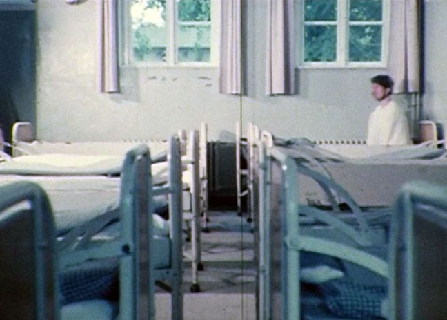 In einem Zimmer, wo viele Bettenin zwei Reihen eng beieinander Stehen, sitzt ein Patient mit Zwangsjacke aufrecht im hintersten rechten