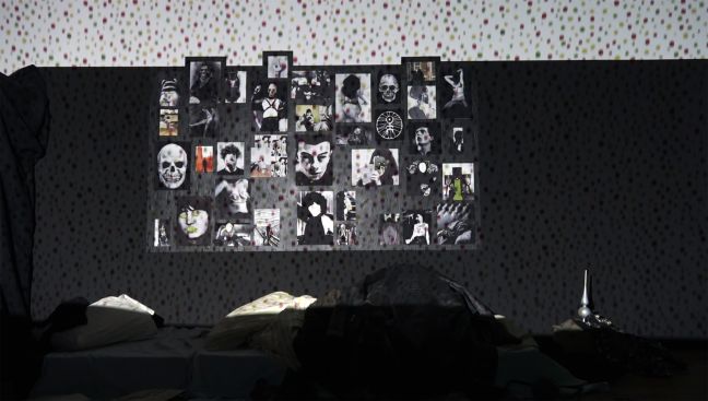 Ein Bett, darüber bunt gepunktete Tapete, ein Großteil schwarz verhängt ein Moodboard an der Wand mit Bildern von Knochenmännern und verfremdeten Detailaufnahmen von Frauen