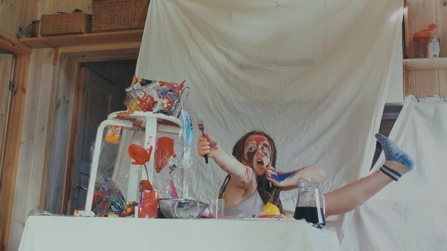 Roosi mit 15 Jahren, spielt ein Schauspiel, sitzt bemalt hinter einem Stapel Plastik in Abwehrhaltung mit einer Gabel am ausgestreckten Arm