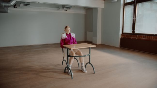 Kielo (Magenta Kostüm) sitzt an ihrem Tisch