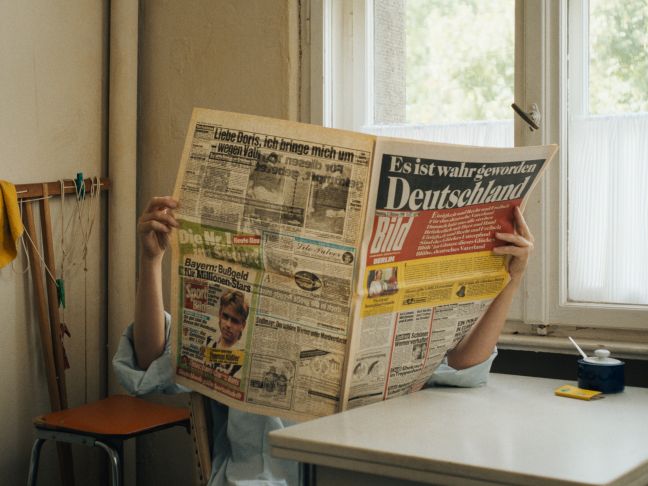 Eine Person sitzt an einem Tisch mit einer aufgeschlagenen Zeitung, in der es um die Wiedervereinigung geht