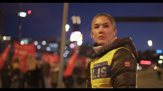 Abends, Dialogpolizistin Nicole steht im Licht der Straßenbeleuchtung, im Hintergrund Demonstranten