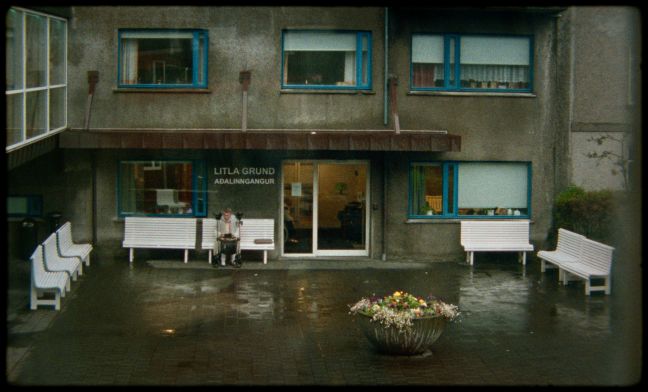 Protagonistin sitzt draußen auf der Bank, vor dem Seniorenheim, Aufnahme vom weitem, Gebäude auch sichtbar