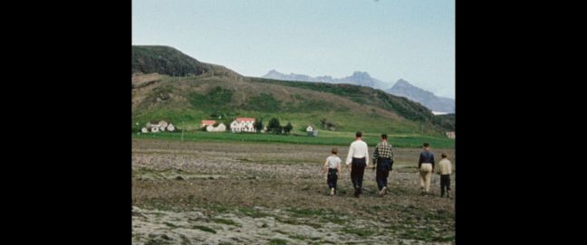 eine Familie geht über ein Feld Richtung Häuser, im Hintergrund Berge